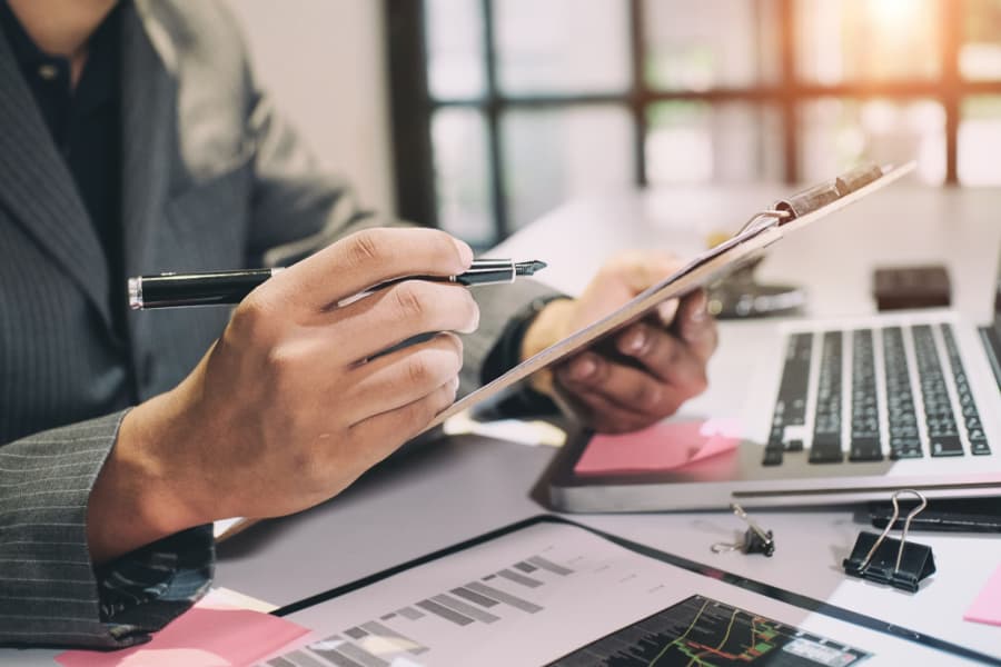 Imagem de uma pessoa escrevendo em um tablet em um escritório com computador e papeladas na mesa como fundo